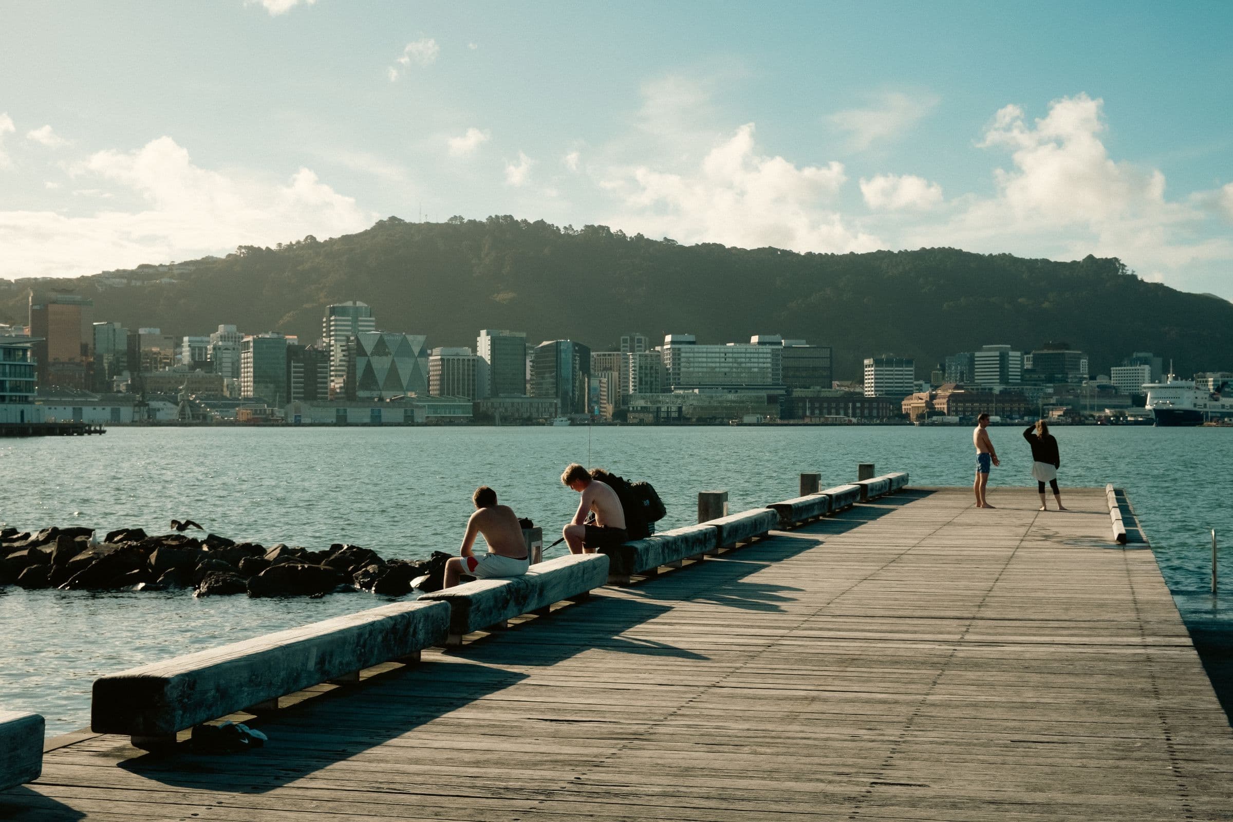 Dermaga kayu di tepi air Wellington pada siang hari, dengan beberapa orang duduk dan berdiri serta garis langit kota dan perbukitan di seberang teluk