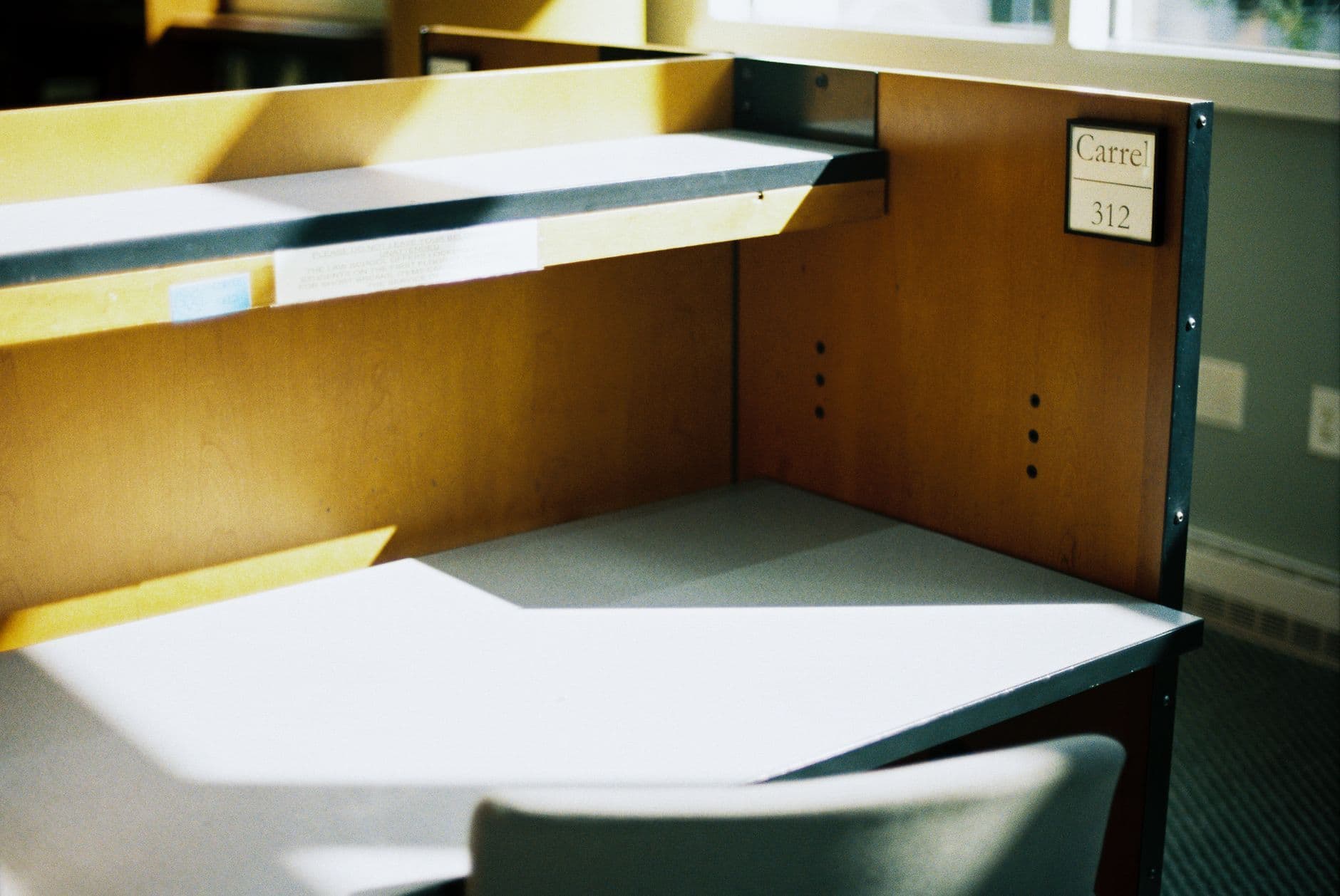 Meja belajar carrel kosong di perpustakaan yang diterpa cahaya matahari dari jendela, tanpa orang