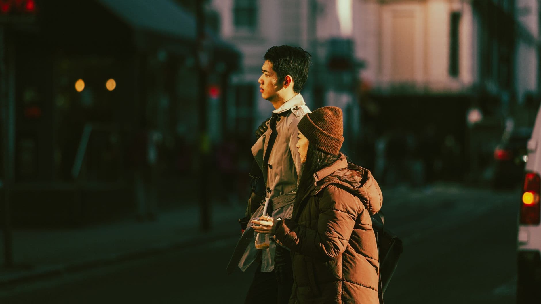 Dua orang dewasa berjaket musim dingin berdiri di tepi jalan kota pada senja, dengan etalase dan lampu kendaraan yang buram di belakang mereka.