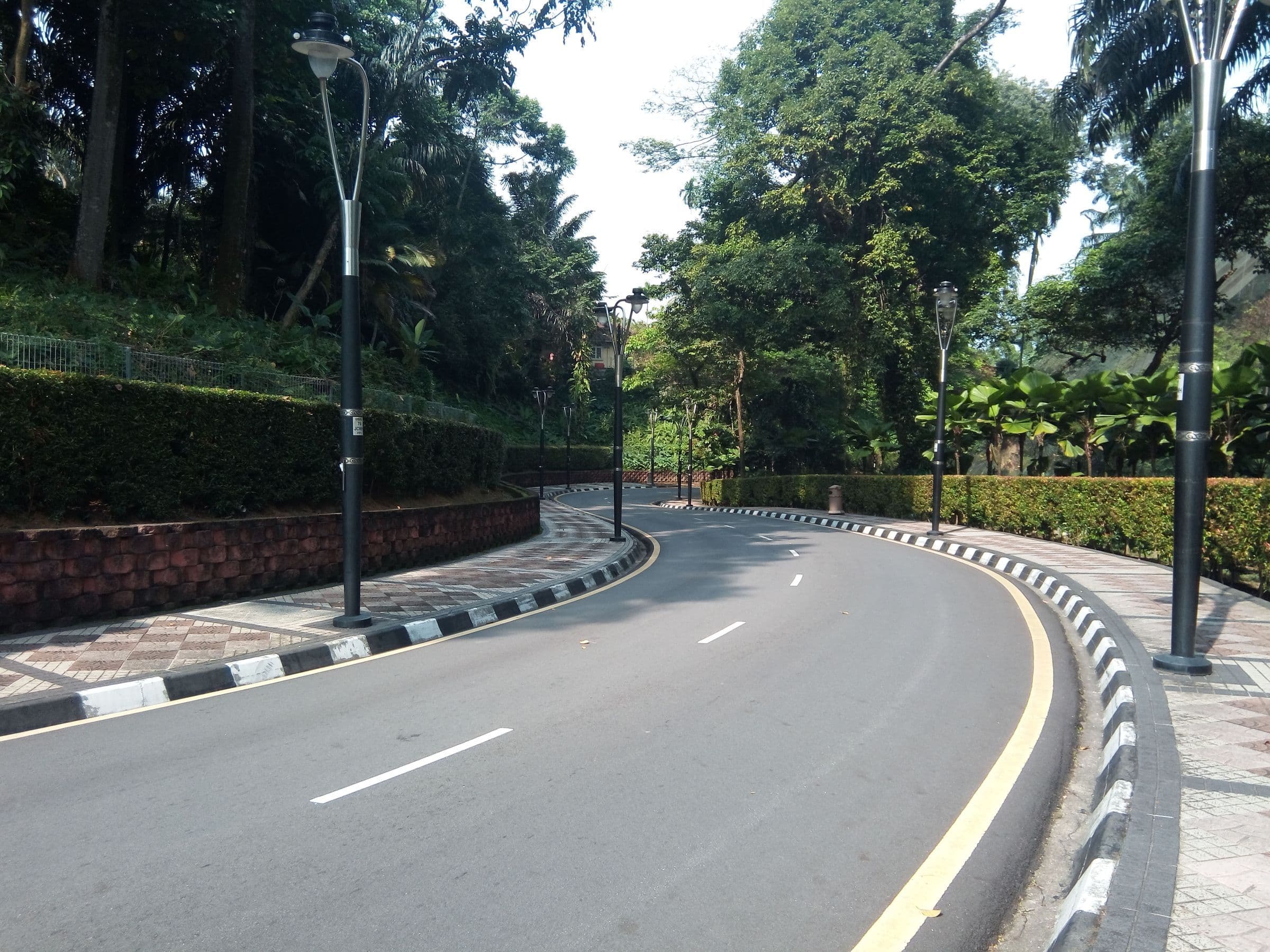 Jalan beraspal yang melengkung di antara pepohonan hijau dan tiang lampu di Kuala Lumpur, tanpa kendaraan dan tanpa orang