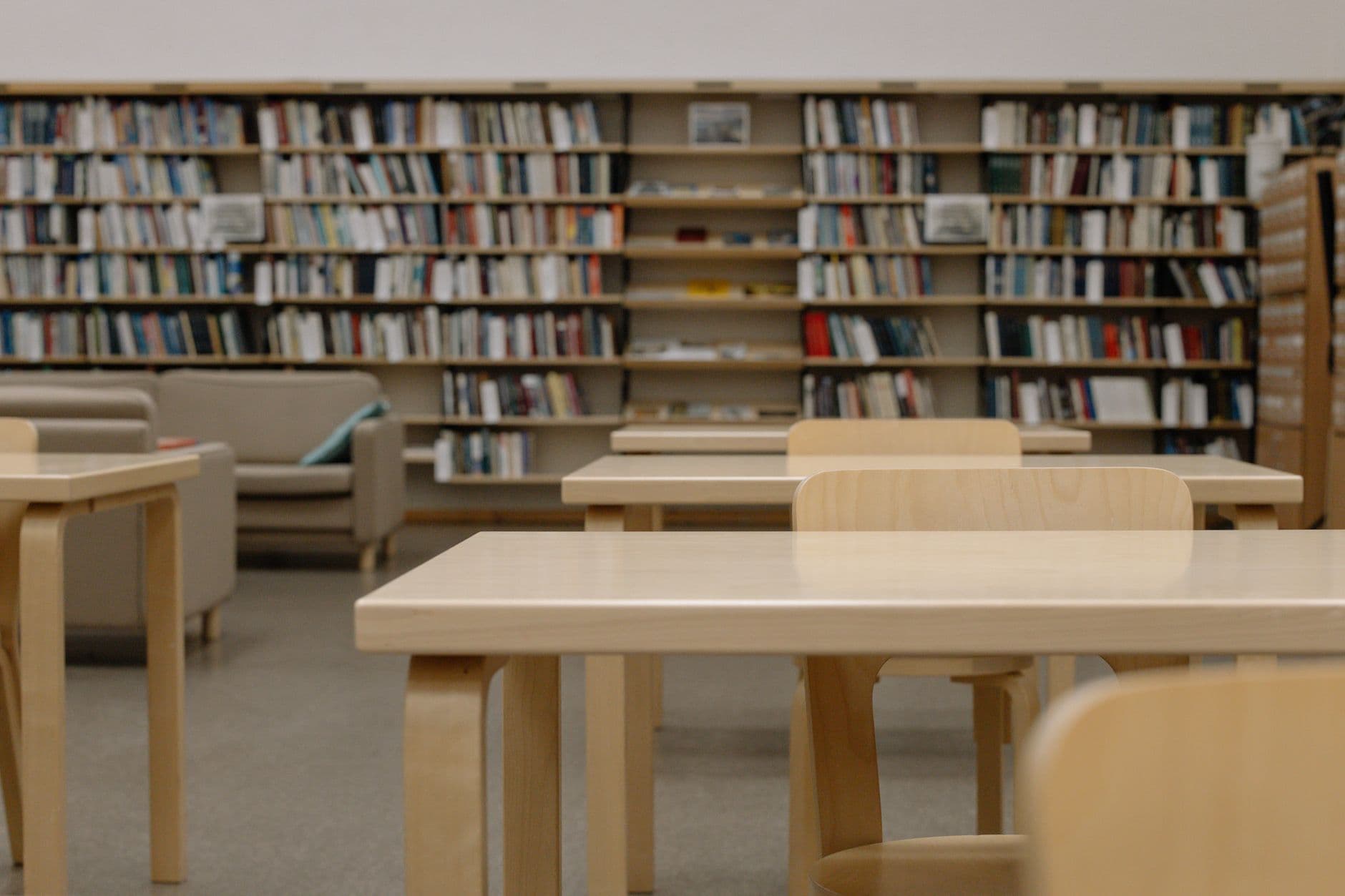 Ruang baca perpustakaan yang kosong dengan meja kayu terang dan rak buku penuh di latar belakang